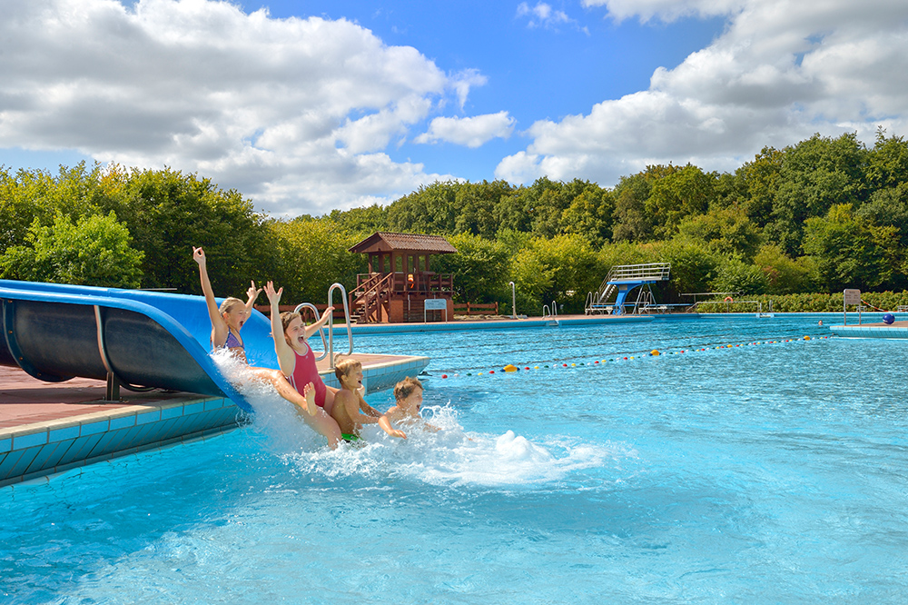 Blick auf das kombinierte Schwimmbecken des Freibad Waldbad Gnarrenburg. Kinder auf der großen Wasserrutsche. Der Sprungturm im Hintergrund.